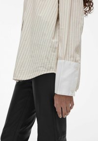 Gestreepte lange mouwen shirt in lichtbeige met subtiele pastelstrepen, met een witte manchet met knopen. Draagt een zwarte leren broek.