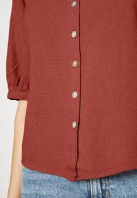 Camisa de cor ferrugem feita de tecido texturizado, com um fecho de botões na frente com seis botões circulares de cor clara e mangas bufantes.