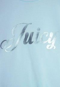 Világoskék t-shirt, amelyen nagyméretű, ezüst fémszínű, dőlt betűkkel olvasható a "Juicy" felirat. Az anyaga simának tűnik, és kereknyakú fazonú.