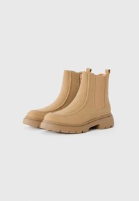 Par de botas de tobillo de ante color beige con suelas de goma gruesa, paneles laterales elásticos y cierres de cremallera sobre un fondo blanco liso.