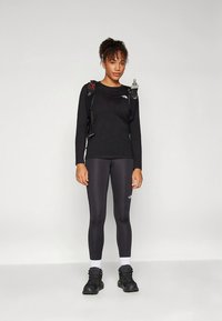 Černé sportovní tričko s dlouhým rukávem, černé legíny a černé boty. Přes rameno je nasazen batoh na hydrataci s připevněnou lahví na vodu.