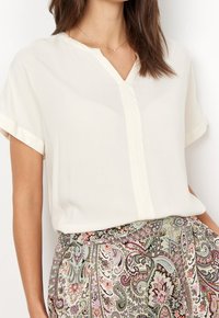 Blouse crème clair avec un col en V, manches courtes retroussées et une texture lisse, associée à une jupe à motifs avec des dessins cachemire.