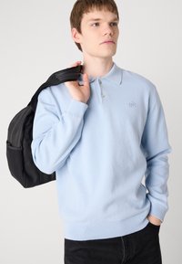 Gaiši zilās krāsas džemperis ar garām piedurknēm un ribinātu apakšmalu. Iekļauj apkakles veida dekoltē un mazu emblemu uz krūts. Melna mugursoma.