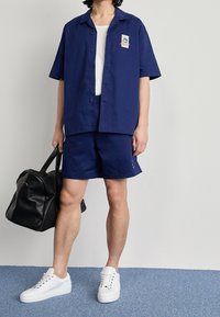 Marinblå kortärmad skjorta med en ficka och logo, i kombination med marinblå shorts. Vita sneakers och en svart väska fulländar looken. Slät tyg, avslappnad passform.