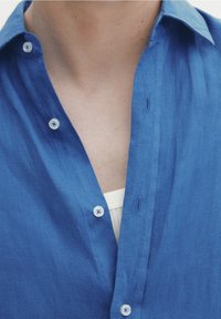 Uppknäppt blå skjorta över vit t-shirt som visar hals och övre bröst.