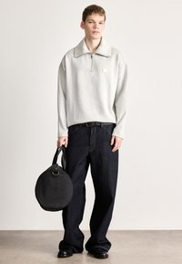 Γκρι ανοιχτό φλις πουλόβερ με φερμουάρ στον γιακά, σε συνδυασμό με σκούρα μπλε άνετο τζιν παντελόνι. Κρατώντας μια στρογγυλή μαύρη τσάντα με πάνω χερούλι.