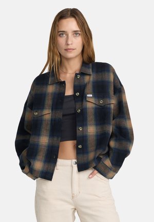 Jonge vrouw draagt een donker ruitjesblouse over een zwart crop top en licht beige broek, staand met handen in de zakken tegen een effen achtergrond.