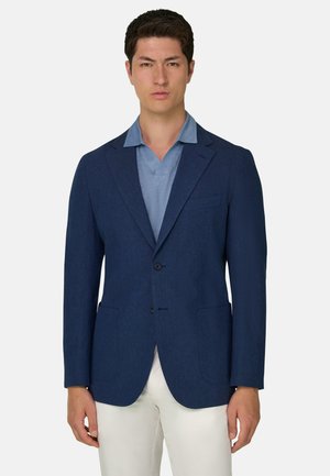 Man draagt een donkerblauw colbert over een lichtblauw overhemd met kraag en witte broek, staand tegen een effen witte achtergrond.