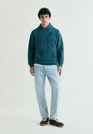 Jeune homme debout, portant un sweat à capuche bleu sarcelle, un jean bleu clair et des baskets marron avec des lacets blancs sur un fond clair uni.