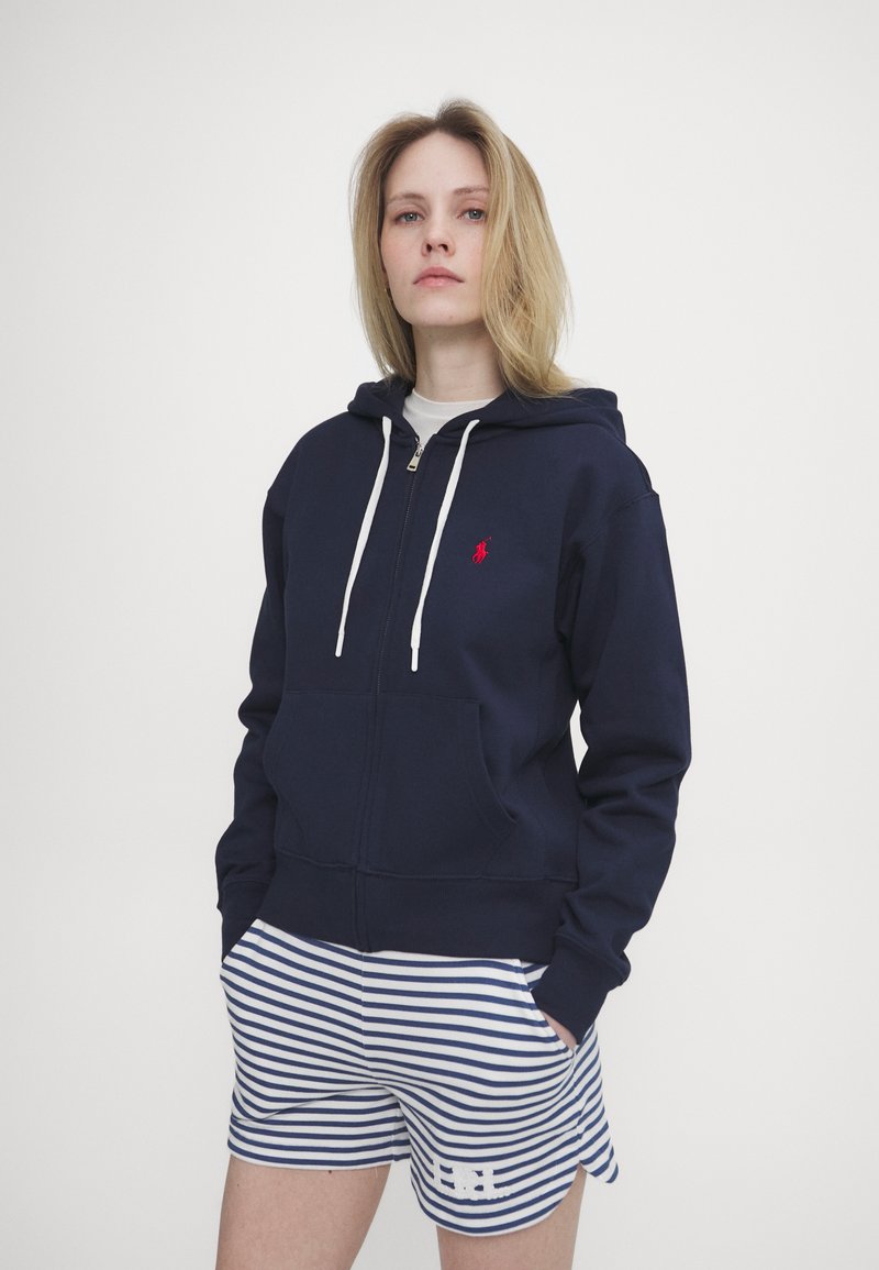 Moleton azul com fecho e drawstrings brancas e um pequeno logótipo vermelho, combinado com calções às riscas azuis e brancas com um detalhe decorativo.