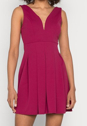 Robe sans manches magenta avec un décolleté en V profond et une jupe plissée portée par une personne aux cheveux bouclés sur un fond uni.