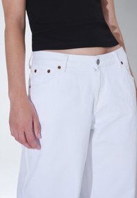 Los jeans de mezclilla blancos presentan un diseño de pierna recta, cinco bolsillos y acentos de hardware metálico en bronce. La tela tiene un acabado texturizado.