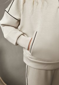 Light beige sweatshirt with black piping accents, featuring a front pocket and ribbed cuffs for texture. Soft fabric with a relaxed fit.