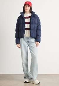 Granatowa kurtka puchowa z wysokim kołnierzem, biała sweter z czerwono-niebieskimi paskami, jasnoniebieskie szerokie dżinsy i białe trampki. W zestawie czerwona czapka.