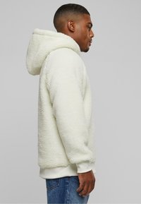 White fleece hoodie with a textured surface, a large hood, and ribbed cuffs, paired with blue jeans. Side view showcases design and fit.