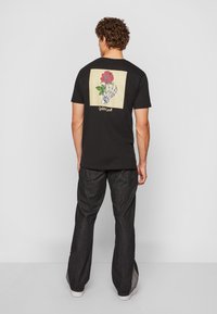 Black t-shirt with a back graphic of a rose held by a skeleton hand, framed in a beige square. Worn with dark denim pants.