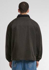 Dark brown jacket with a high collar, elastic cuffs, and a relaxed fit. Notable embroidered accents on back pockets.
