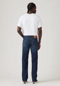 Jeans de mezclilla azul oscuro con pierna recta, que cuentan con dos bolsillos traseros, un parche de marca y costuras visibles. Llevados con una camiseta blanca.