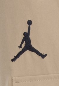 Beige stof met een donkerblauwe geborduurde silhouet van een springende figuur die een basketbal vasthoudt. Zakdetail zichtbaar op de stof.