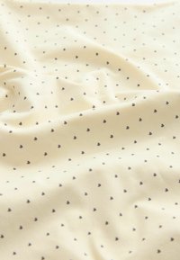 Tissu de couleur crème avec de petits motifs de cœurs noirs uniformément espacés, présenté avec des plis doux et des rides.