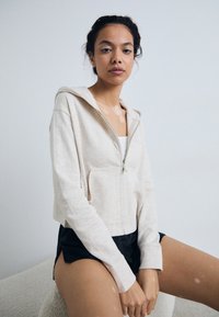 Cropped, hoodie com fecho éclair em tecido bege claro, apresentando capuz e bolsos frontais, combinado com calções pretos. Textura suave e ajuste descontraído.
