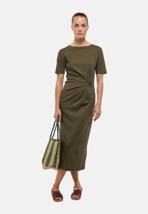 Femme portant une robe midi olive à manches courtes avec torsade latérale, portant un sac tissé rayé vert et beige, et des sandales bordeaux.