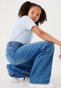 Jeans de mezclilla azul de pierna ancha con una cintura alta, que presentan un parche de cuero en la parte trasera y detalles de costura sutiles, combinados con una camiseta azul clara.