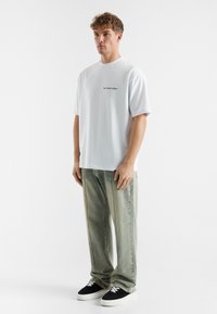 Hvit t-skjorte med korte ermer og svart tekst, sammen med løstsittende lysegrønne jeans og svarte joggesko med hvite snører.