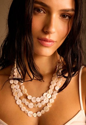 Nuori nainen, jolla on märät tummat hiukset, pukeutuneena kerroksittain valkoisiin pyöreisiin simpukkakaulakoruihin ja beigeen hihattomaan topiin neutraalia taustaa vasten.