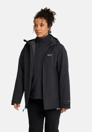 Vrouw met krullend haar, die een zwarte Jack Wolfskin capuchonjas en bijpassende zwarte broek draagt, staand en licht naar de zijkant kijkend.