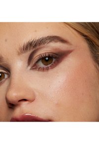 Gros plan sur un œil mettant en valeur un fard à paupières foncé avec un motif angulaire, des cils longs et un teint de peau lisse. Les lèvres sont brillantes et de couleur neutre.