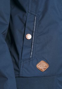 Marinblå jacka med textilstruktur, ficka med dragkedja och en bronssymbol med soldesign. Ribbad manschett fulländar designen.