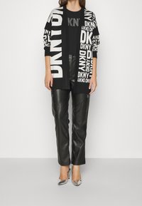 Svartvit cardigan med stort "DKNY"-textmönster, tillsammans med svarta läderbyxor. Modellen bär metalliska klackar som visar upp en elegant design.