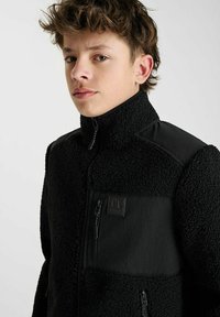 Zwarte fleece jas met hoge kraag, voorzien van textuurmateriaal, een voorzak met rits en een soepel synthetisch paneel op de borst.