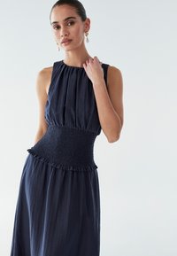 Abito senza maniche di un blu navy scuro con tessuto testurizzato, vita arricciata e dettagli orlo arricciato. La modella posa con una mano sulla spalla, mostrando il design.