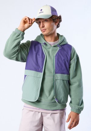 Junger Mann, der eine weiße und lila Mütze zurechtrückt, trägt einen hellgrünen Fleece-Hoodie mit lila Weste und hellfarbige Hose.