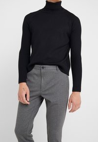 Man in een zwarte geribbelde coltrui en grijze geknoopte broek, staand tegen een effen lichte achtergrond.