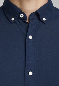 Camisa azul marino abotonada con cuello clásico, con botones blancos y un tejido texturizado. Costuras visibles a lo largo de los bordes.