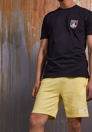 Młoda osoba ubrana w czarną koszulkę ellesse i żółte szorty stoi na tle zużytej, poplamionej drewnianej ściany.