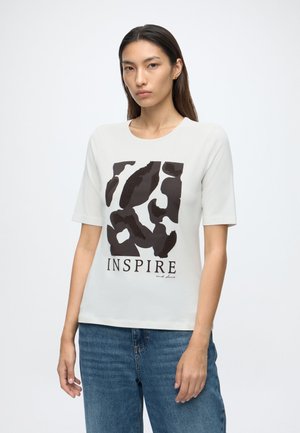 Młoda kobieta z długimi ciemnymi włosami, ubrana w białą koszulkę z czarnymi abstrakcyjnymi kształtami i słowem "INSPIRE", w połączeniu z niebieskimi dżinsami.