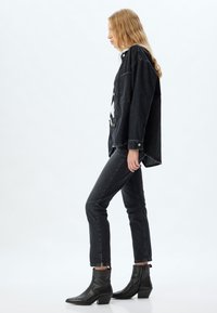 Chaqueta de mezclilla negra con botones a presión, combinada con jeans negros de cintura alta y botines negros. La modelo está de lado contra un fondo claro.