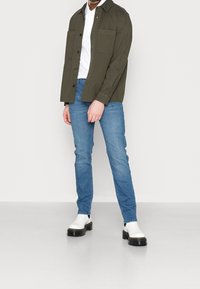 Man med en olivgrön skjorta med knappar över en vit polo, blå slim fit jeans och vita klumpiga stövlar som står mot en enfärgad bakgrund.