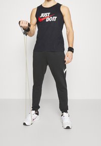 Man in een zwart Nike tanktop en broek die een weerstandsband gebruikt voor een arm oefening, met witte Nike sneakers en een polsband.