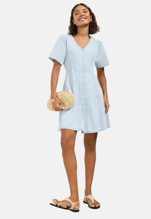 Nő világoskék, gombos, rövid ujjú ruhában, bézs színű fonott kézitáskát tart, és bézs lapos szandált visel.