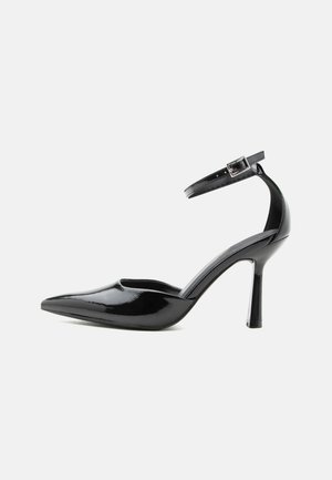 Zapato de tacón alto de charol negro con punta puntiaguda, correa en el tobillo y hebilla plateada, tacón de aguja, vista lateral sobre fondo liso.