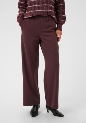 Personne portant un pantalon ample violet foncé, un pull assorti à motifs, et des bottines noires à talons pointues, se tenant devant un fond uni.