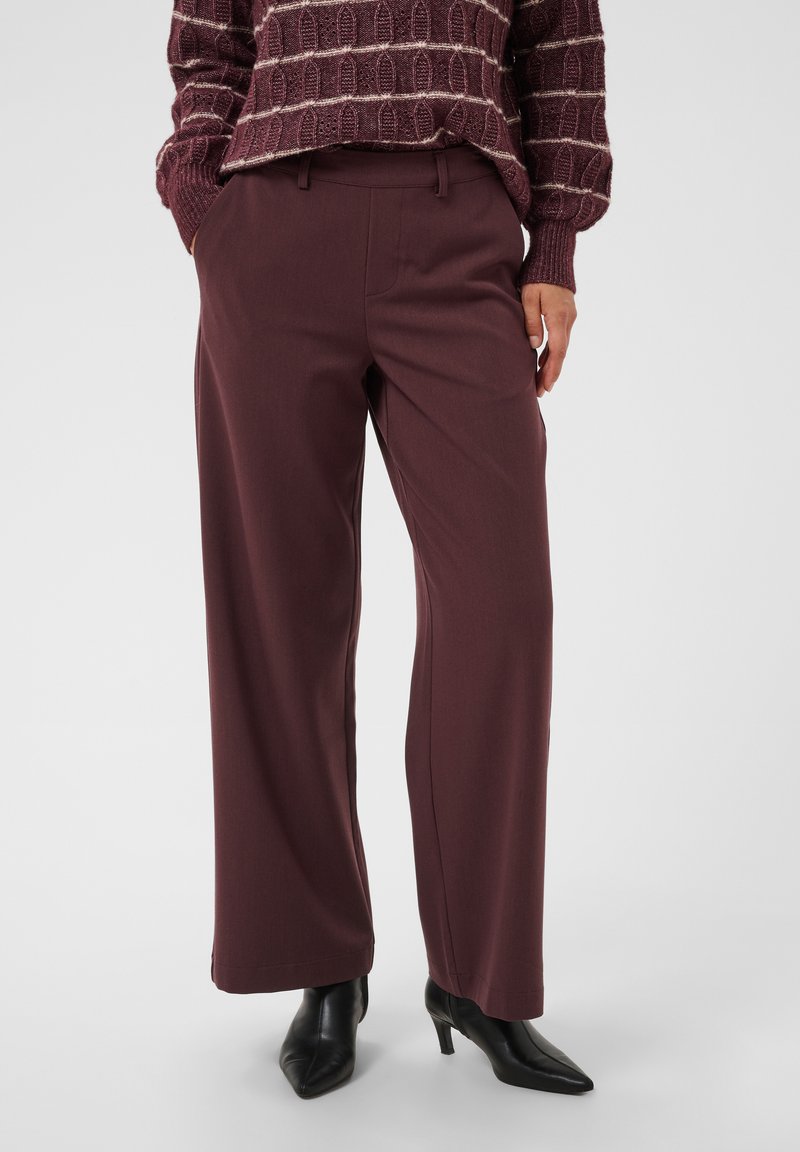 Personne portant un pantalon ample violet foncé, un pull assorti à motifs, et des bottines noires à talons pointues, se tenant devant un fond uni.