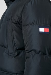 Černá prošívaná péřová bunda s prošívaným vzorem. Má vodorovnou kapsu, žebrované manžety a na rukávu nášivku s logem v červené, bílé a modré barvě.