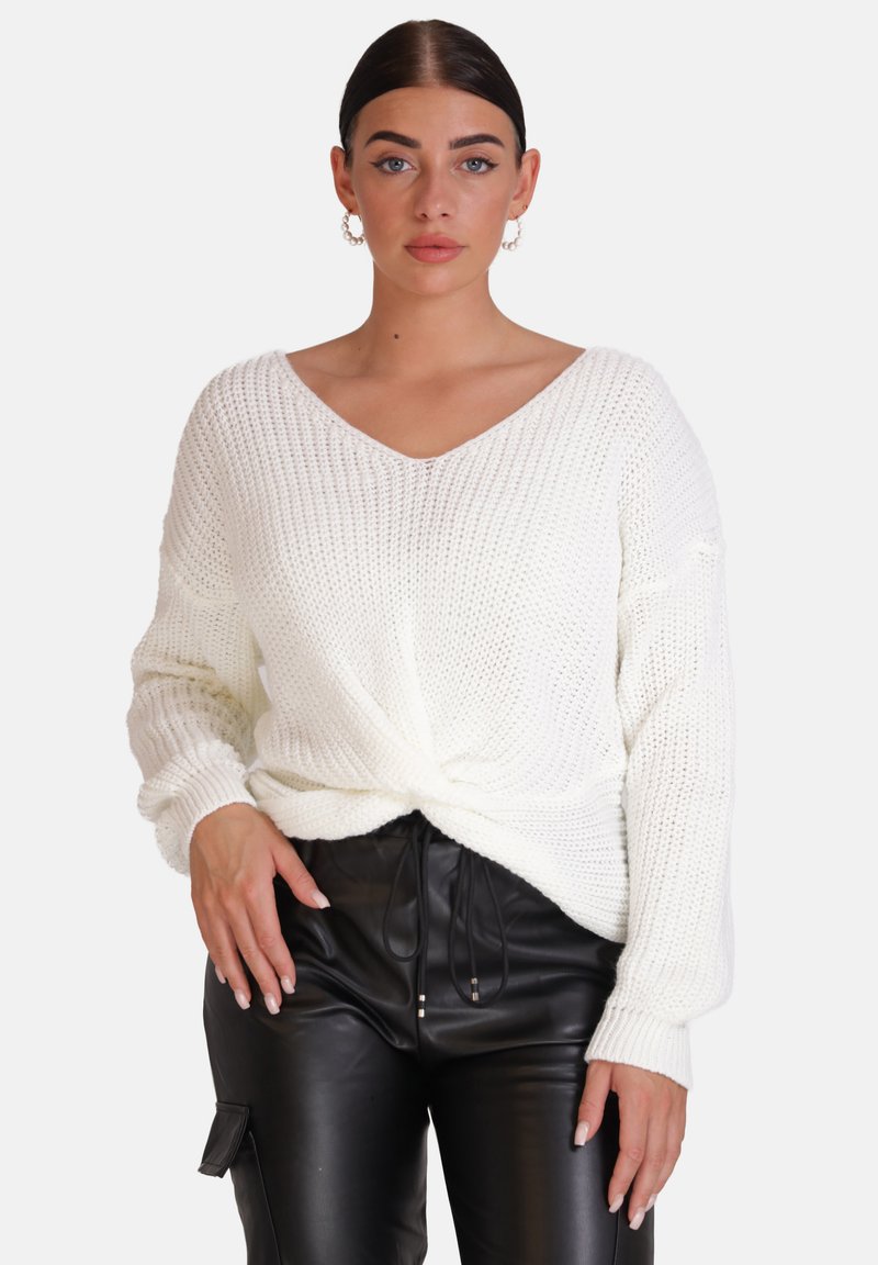 Weißer, gestrickter Pullover mit V-Ausschnitt, lockerer Passform und gedrehtem Front-Detail, kombiniert mit schwarzen, lackierten Kunstlederhosen.