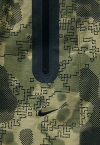 Groene camouflage stof met een textuurpatroon en zwarte accenten, een rits aan de voorkant en een klein Nike-logo onderaan.